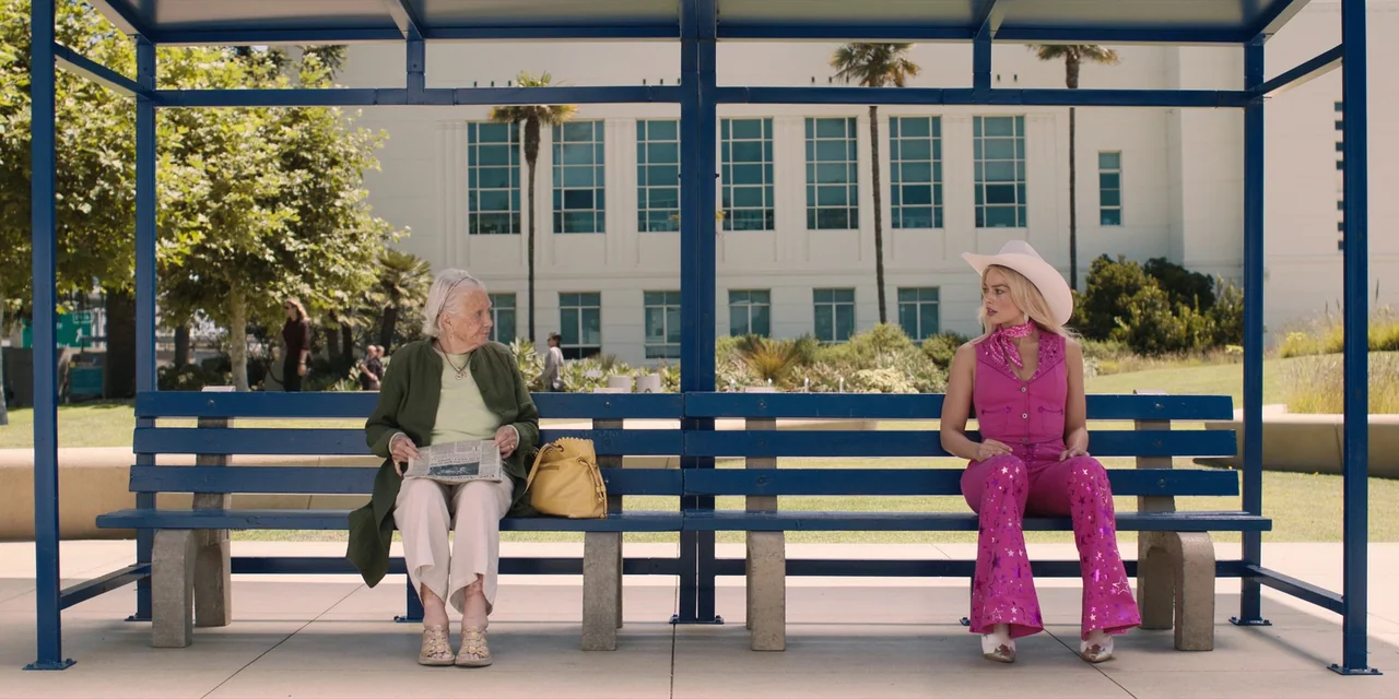 Barbie (2023) movie still: wide — A sunny public bus stop features a blue metal bench and white building facade. An elderly woman reads a newspaper…