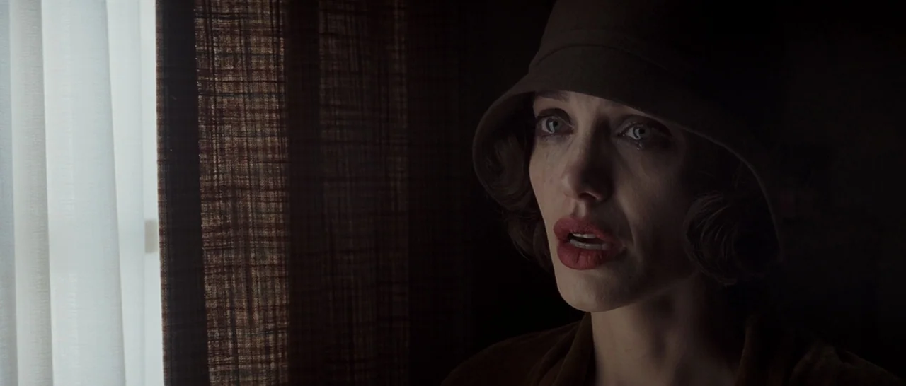 Changeling (2008) movie still: closeup — A dimly lit room with textured dark curtains hanging beside a bright window. A woman wearing a 1920s-style…
