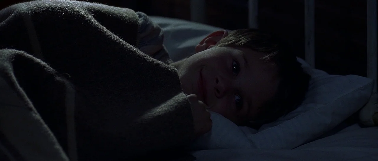 The Cider House Rules movie still: closeup — A dark bedroom shows a child lying in bed at night. The boy rests his head on a white pillow while…