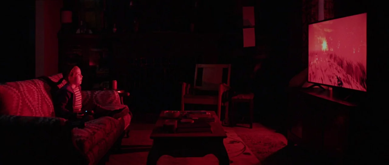 The Boogeyman (2023) movie still: wide — A dark room features a patterned sofa, a wooden coffee table, and a television. A child sits on the couch…
