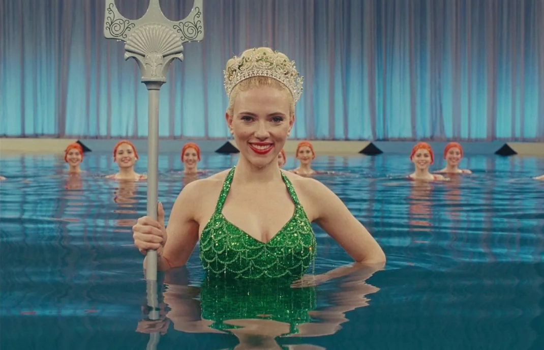 Hail, Caesar! (2016) movie still: medium — A woman stands in a brightly lit swimming pool backed by blue draped curtains. Other women in swim caps…