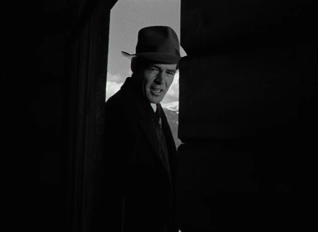 On Dangerous Ground (1951) movie still: medium — A man stands in the threshold of a dark wooden building against a bright background. He peers out…