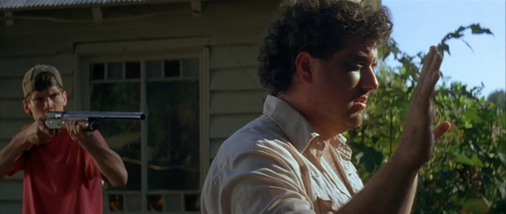 Shotgun Stories (2007) movie still: medium — A man stands in front of a wooden house with a window and siding. A teenager holds a shotgun pointed…