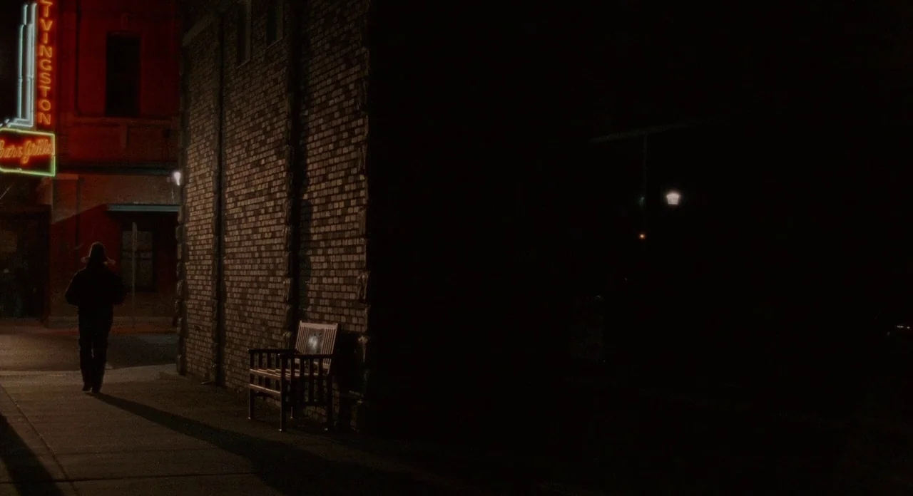 Certain Women (2016) movie still: wide — A dark city street corner at night features a brick building wall and a bench. A lone person in dark…