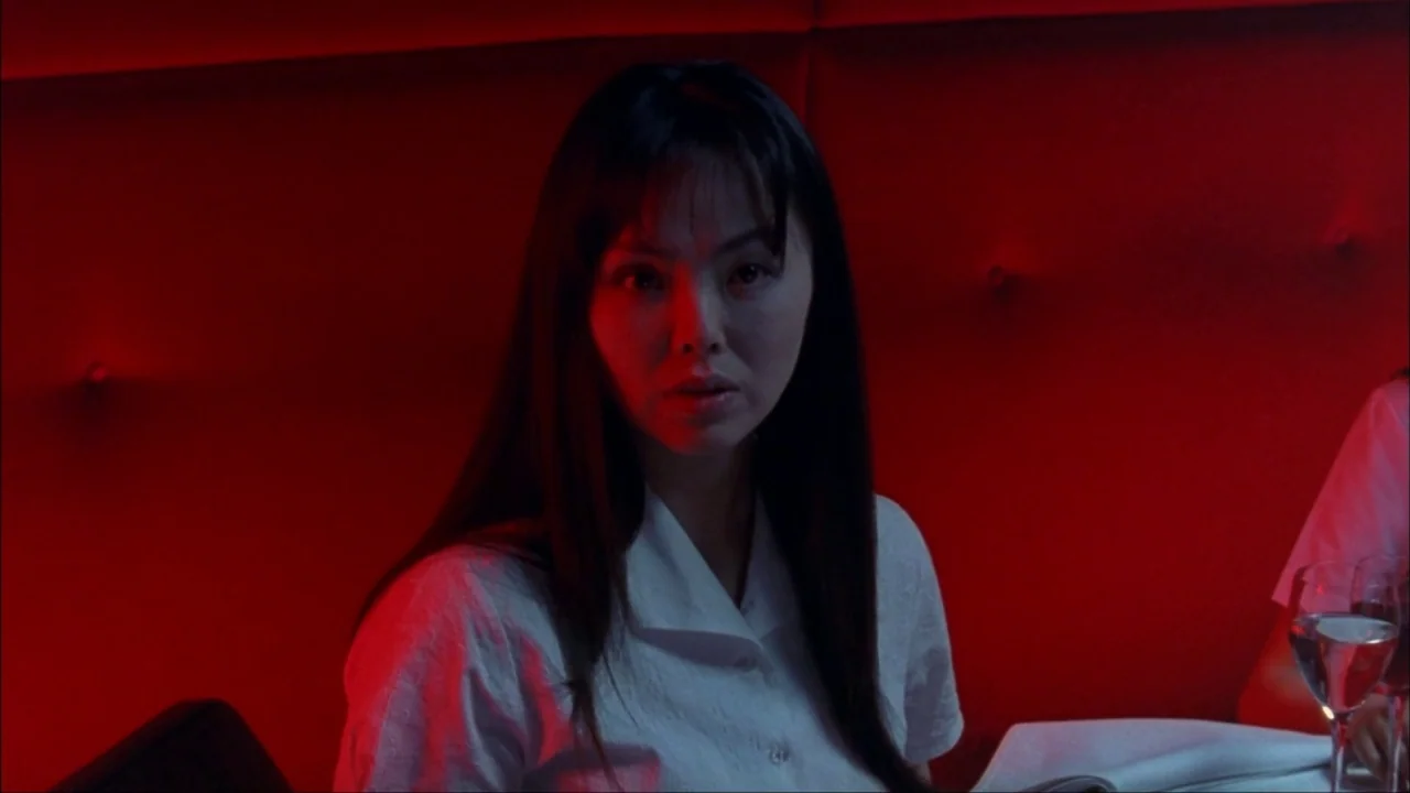 Audition (1999) movie still: medium — A woman sits in a booth with a tufted red backrest. She is looking directly forward.