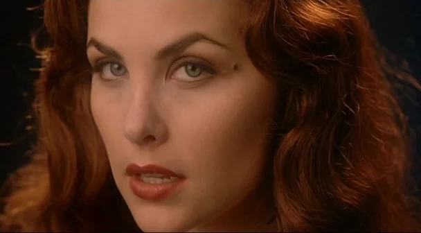 Boxing Helena (1993) movie still: closeup — A close-up shot of a woman with auburn hair against a dark background. She maintains direct eye contact…