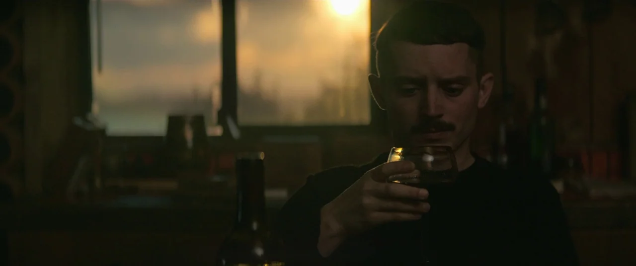 Come To Daddy (2019) movie still: medium — A dimly lit bar setting with a wooden counter and large glass windows. A man sits alone holding a wine…