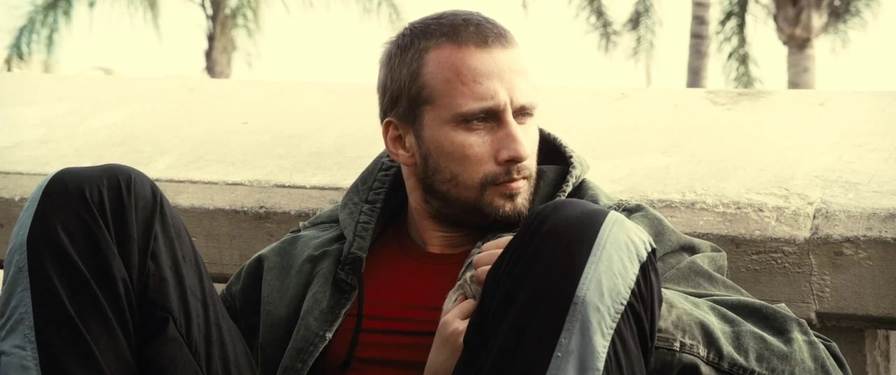Rust And Bone (2012) movie still: medium — A man sits against a concrete wall with palm trees visible in the distance. He wears a layered jacket and…