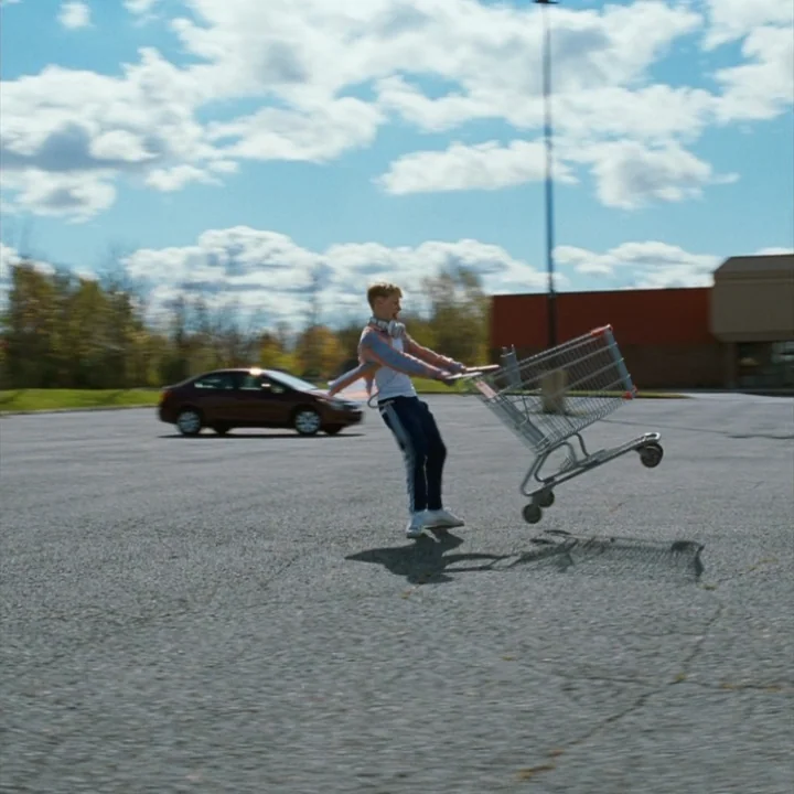 Mommy (2014) movie still: fullbody — A spacious paved parking lot features a distant red car under a blue sky. A teenage boy holds onto a shopping…