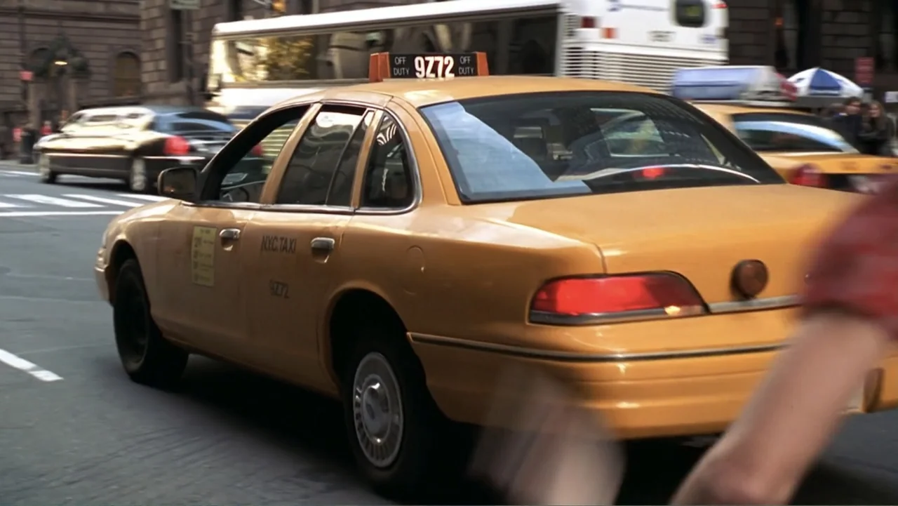 13 Going on 30 (2004) movie still: medium — A yellow taxi cab drives across a paved urban intersection during the day. Other vehicles and a city bus…
