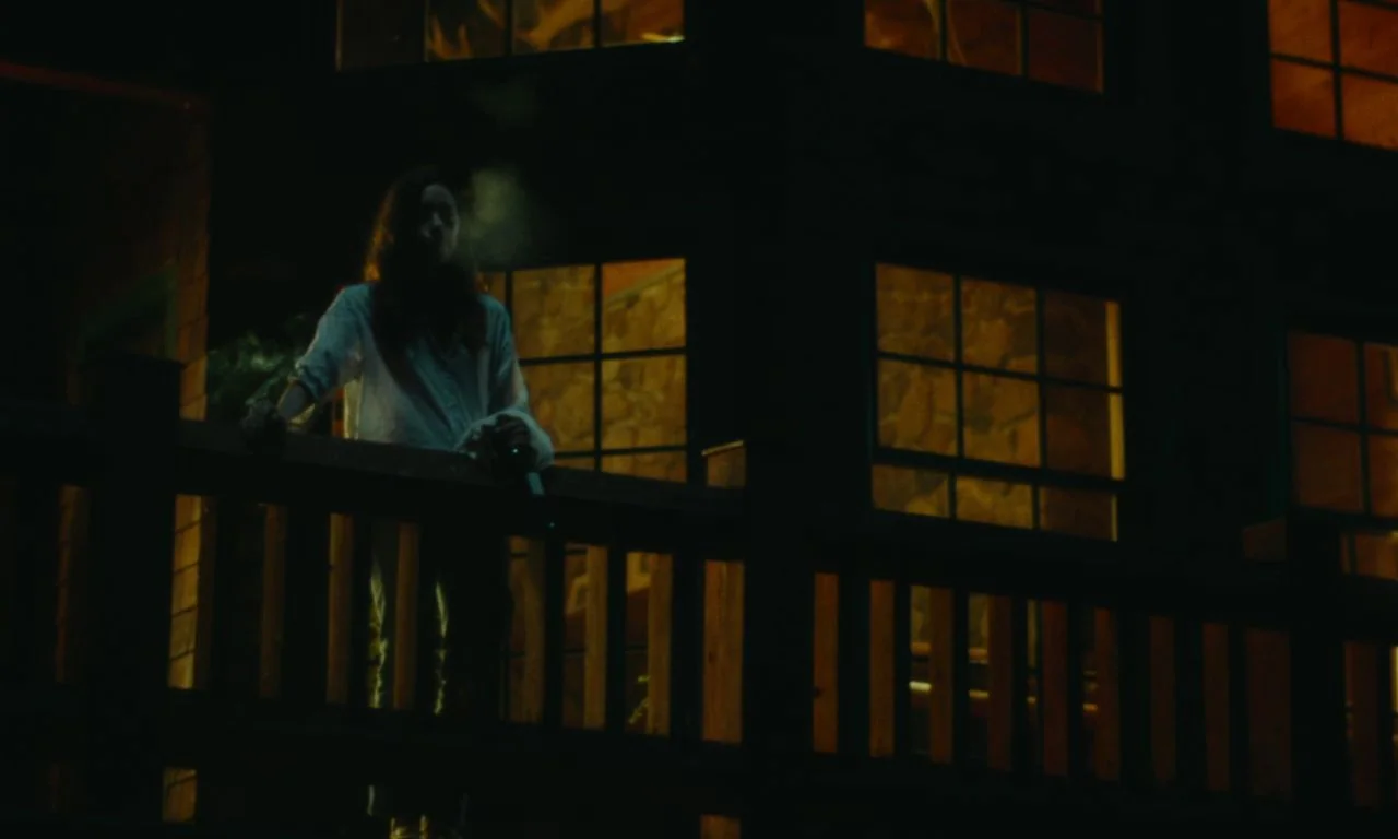 Black Bear (2020) movie still: medium — A woman stands on a wooden balcony at night against a large house facade. The interior of the house glows…