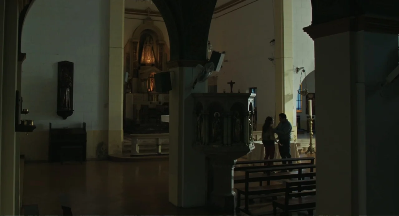 Belén (2025) movie still: wide — A quiet church interior features stone arches, wooden pews, and an ornate altar area. A man and a woman stand…