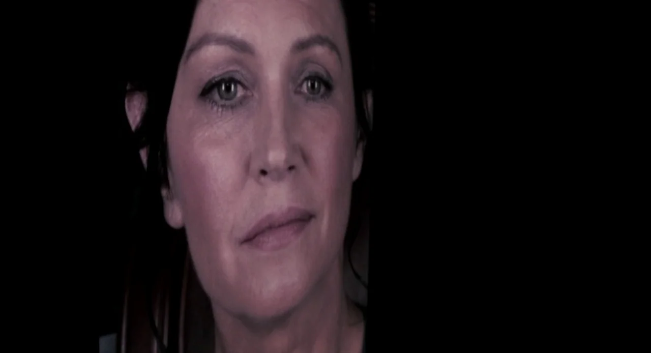 Antiviral (2012) movie still: closeup — A woman's face is positioned in a dark room with minimal background detail. She turns her head slightly…
