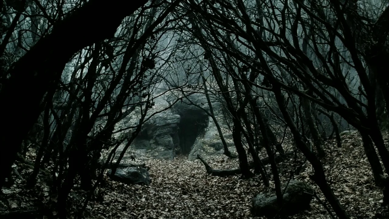 Sleepy Hollow (1999) movie still: establishing — A dark cave opening is framed by skeletal trees and fallen dry leaves. The dense woodland setting…