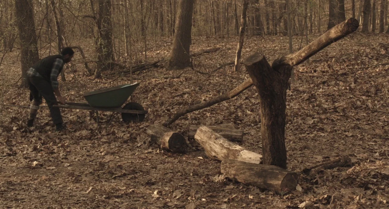 The Alchemist Cookbook (2016) movie still: wide — A dense woodland floor is covered in layers of fallen brown autumn leaves. A man in work clothes…