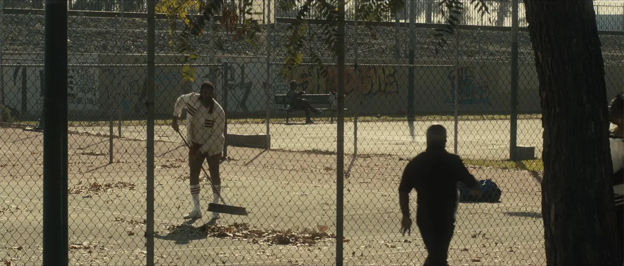 King Richard (2021) movie still: wide — An outdoor fenced tennis court features weathered pavement covered with scattered dry brown leaves. A man in…