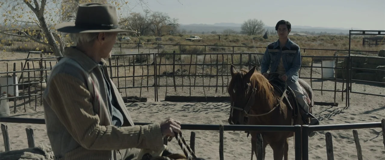 Cry Macho (2021) movie still: medium — A dusty outdoor corral with wooden fence posts under a clear daytime sky. An older man on horseback interacts…