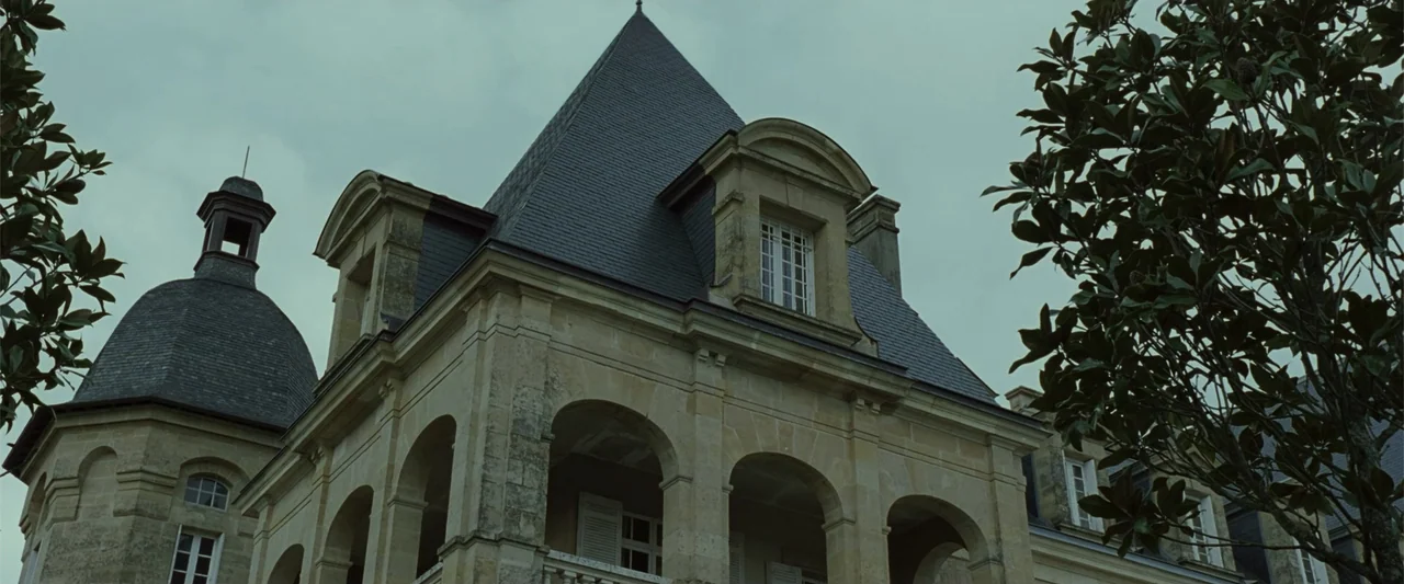 Anna (2013) movie still: establishing — A stone mansion with dark slate roofs and arched columns stands under a sky. A large tree with thick green…
