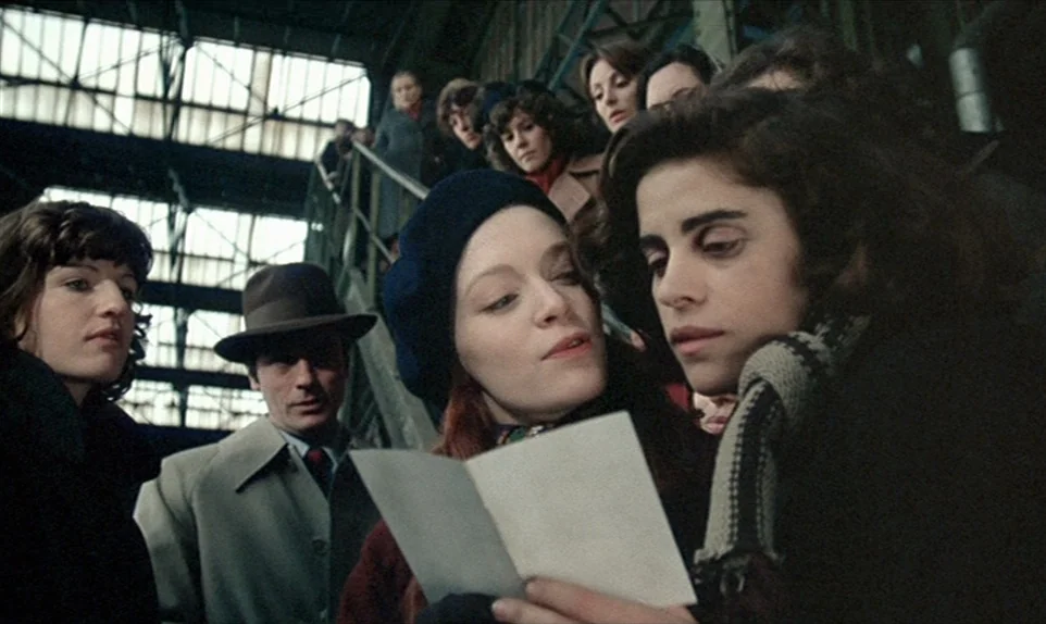 Mr. Klein (1976) movie still: medium — A bustling interior of a train station with an industrial iron ceiling structure. A small group of young…
