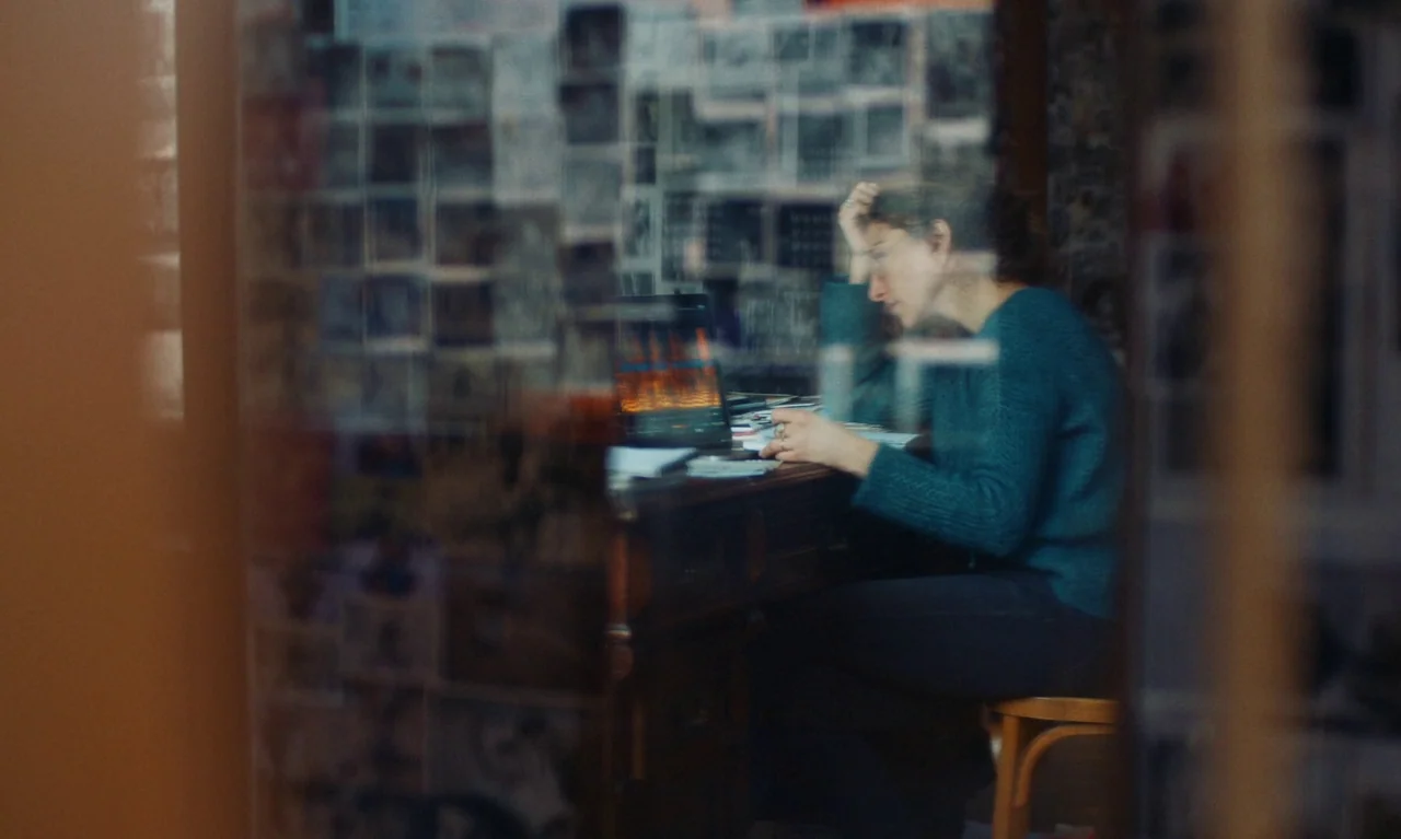 Little Girl Blue (2023) movie still: medium — A person sits at a wooden desk cluttered with papers in a dark room. The view is partially obscured by…