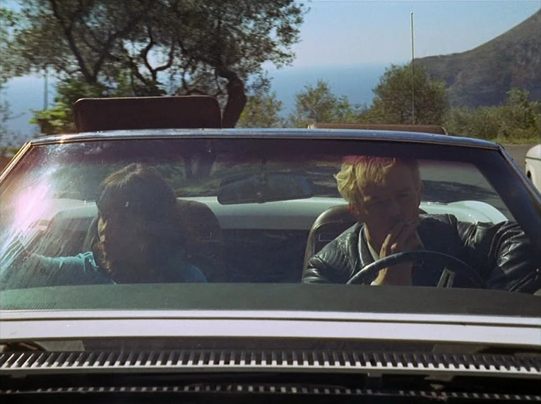Beware Of A Holy Whore (1971) movie still: medium — A convertible car drives along a coastal road with trees visible in the background. A blonde man…