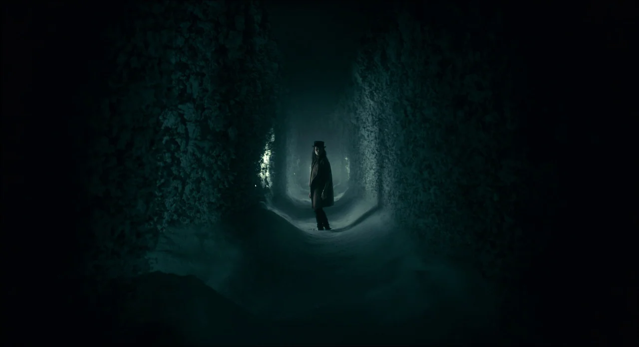 Doctor Sleep movie still: wide — A narrow snow-covered path winds through tall dense hedges in a darkened maze. A woman in a top hat and coat stands…