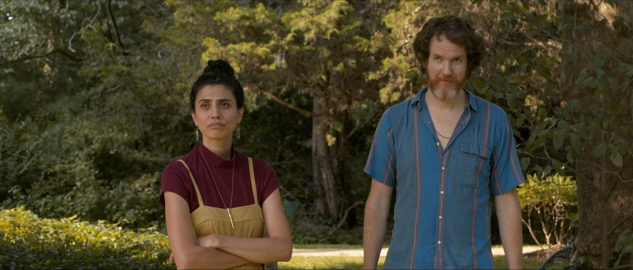 Songs For A Sloth (2021) movie still: medium — An outdoor area with dense green foliage and trees provides a natural background. A man and a woman…
