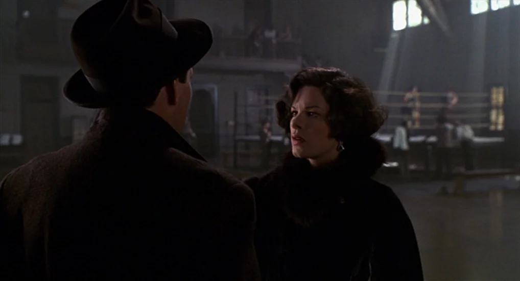 Miller’s Crossing (1990) movie still: medium — An interior space features a boxing ring blurred in the background under high windows. A man in a…