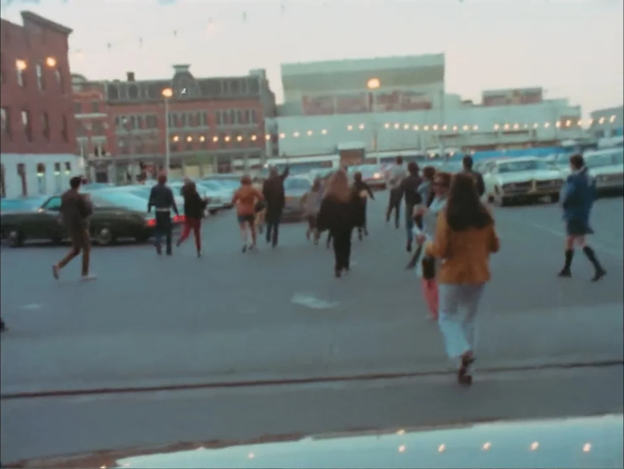 One to One: John & Yoko (2025) movie still: wide — A 1970s outdoor parking lot features several parked vehicles and string lights overhead. A small…
