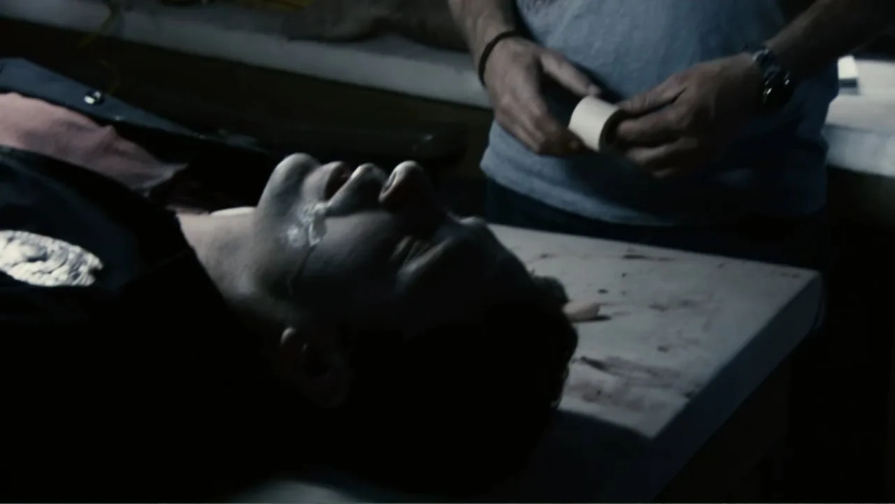 Quarantine (2008) movie still: medium — An interior sterile room with a stainless steel surface holding a person. A standing man wraps medical tape…