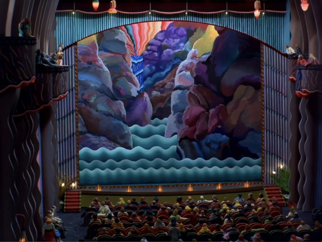 Asparagus (2007) movie still: wide — An Art Deco theater features a large, colorful painted mural of mountains and water. Numerous miniature figures…