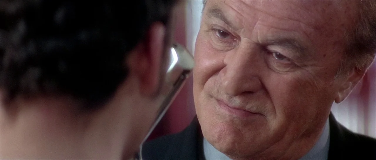 Lost Highway (1997) movie still: closeup — An indoor office setting with a blurred foreground character. The older man stares directly at the…