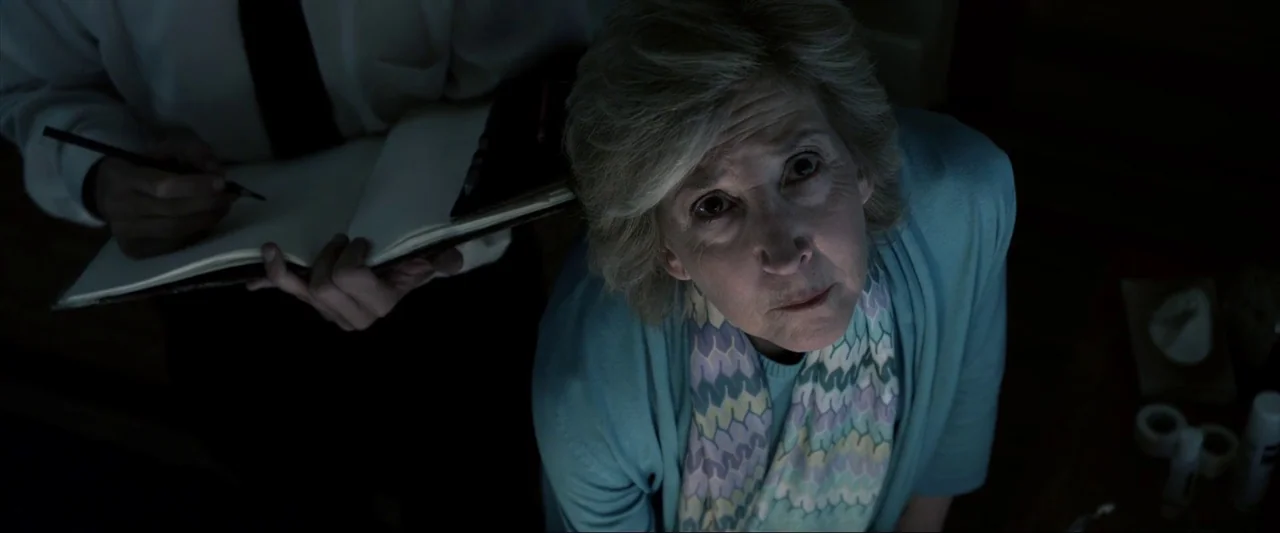 Insidious (2010) movie still: medium — A dimly lit room with blue light catching the textures of clothing and skin. An elderly woman gazes upwards…