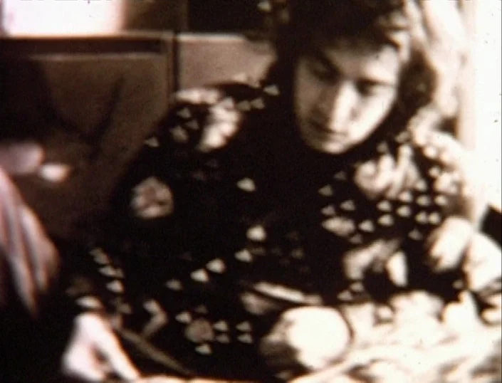 Glitterbug (1994) movie still: medium — A dimly lit room with soft, textured surfaces and low contrast. A young man with long hair looks down while…