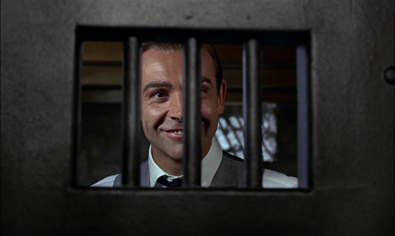 Goldfinger (1964) movie still: closeup — A man looks through the rectangular opening of a heavy iron barred door. His face is framed by the vertical…