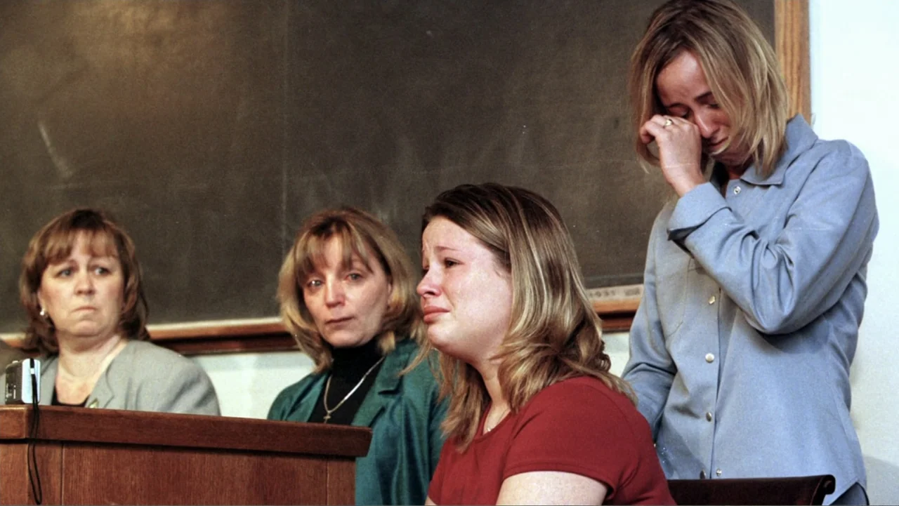 What Happened at Fells Acres? (2023) movie still: medium — A wooden witness stand and dark chalkboard provide the backdrop in a muted room. Four…