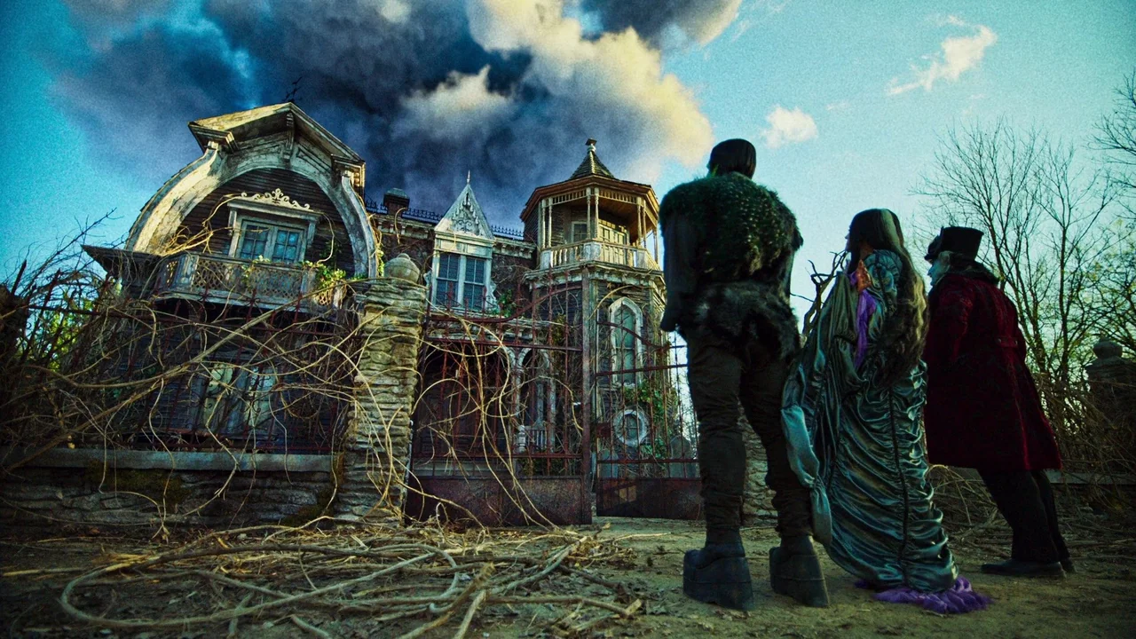The Munsters (2022) movie still: wide — A gothic-style mansion with intricate wood carvings and balconies sits behind a metal fence. Three figures…