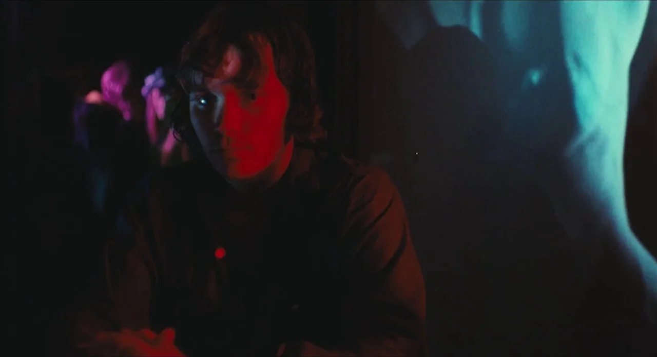 The Other Side of the Wind (2018) movie still: medium — A dark, crowded nightclub with shifting colored lights illuminating the space. A young man…