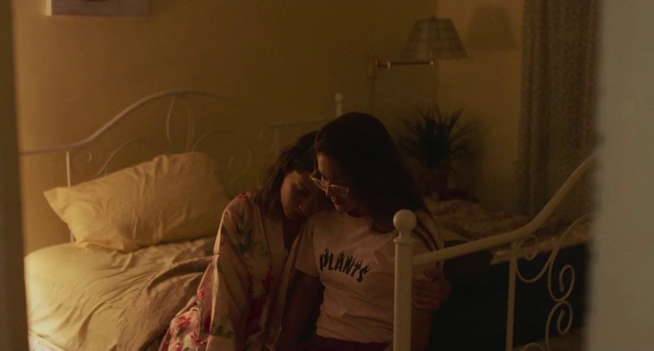 Skate Kitchen (2018) movie still: medium — A dimly lit room with a metal frame bed and floral-patterned bedding. Two young women sit closely on the…