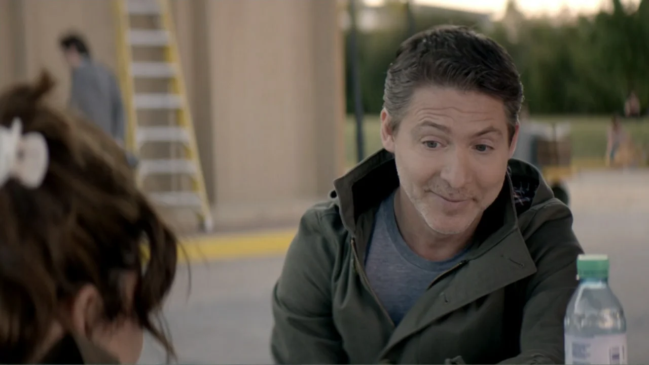 I Married a Dumbass (2016) movie still: medium — A paved outdoor space features a yellow ladder and blurred trees in the background. A middle-aged…