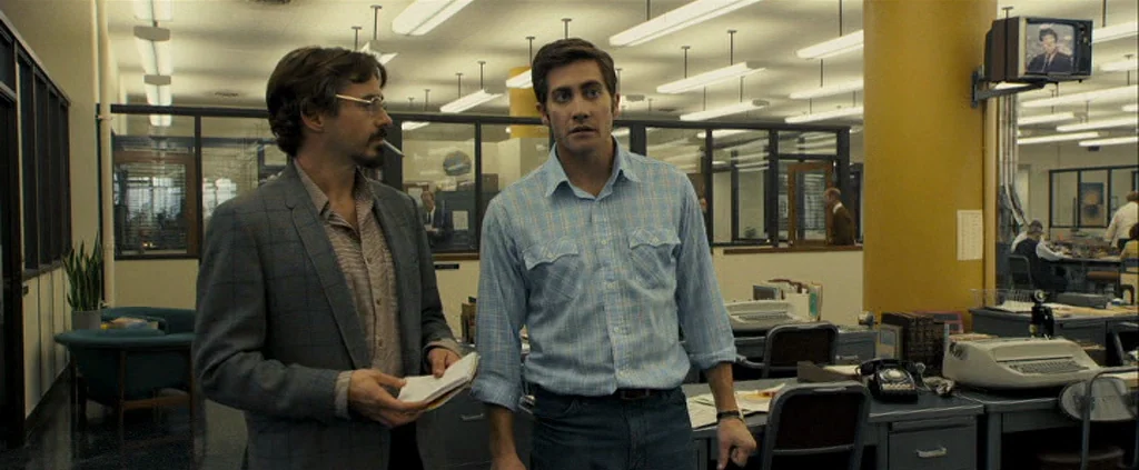 Zodiac (2007) movie still: medium — A 1970s newspaper office with rows of desks and glass partitions. Two men stand in the foreground having a…