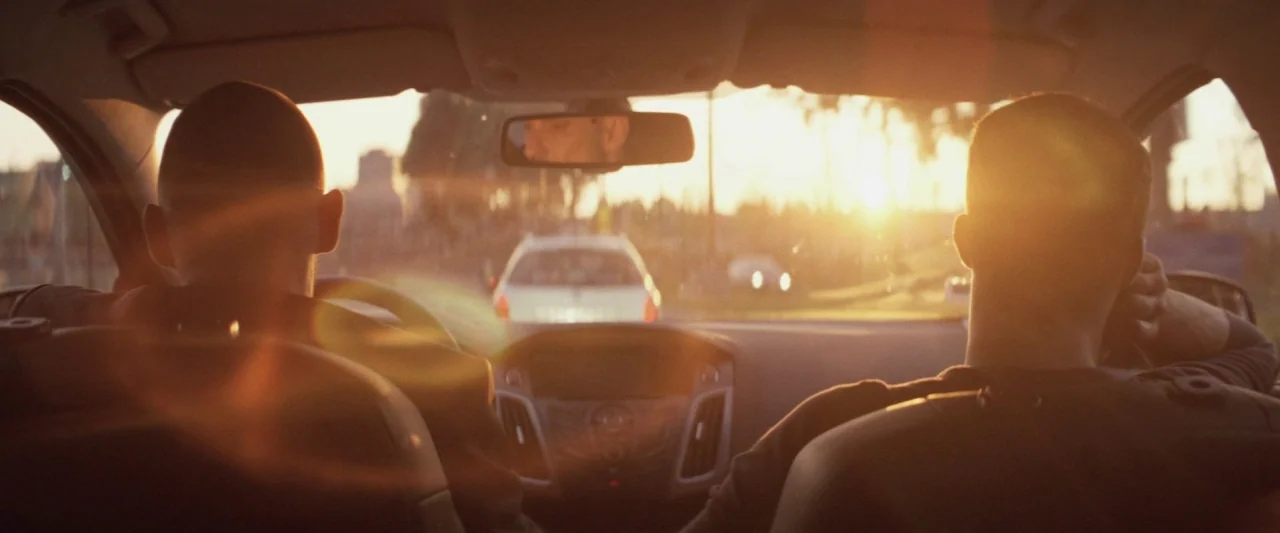 Colt 45 (2014) movie still: medium — The interior of a car during sunset with two men sitting in front seats. A road stretches out ahead through the…