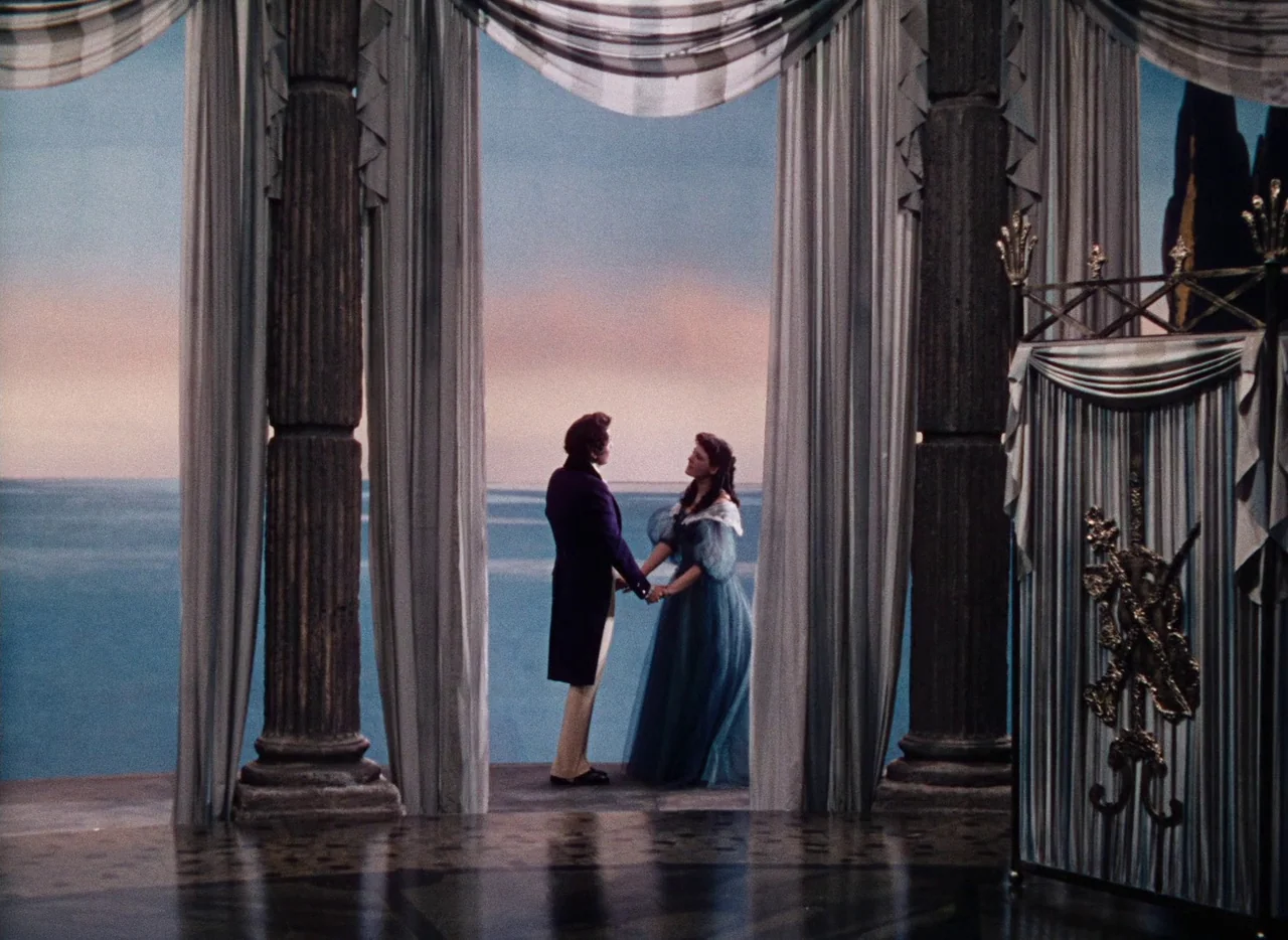 The Tales of Hoffmann (1951) movie still: fullbody — A stone balcony overlooking the sea at sunset framed by large draped curtains. A man and a…