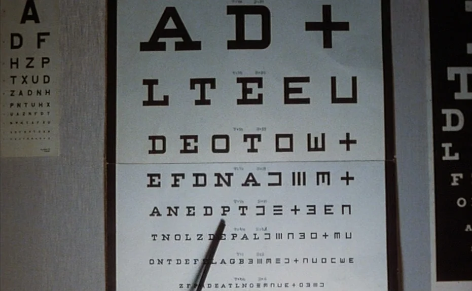 Soldier of Orange (1977) movie still: closeup — A white wall displays several printed eye examination charts with various letters and symbols. A…