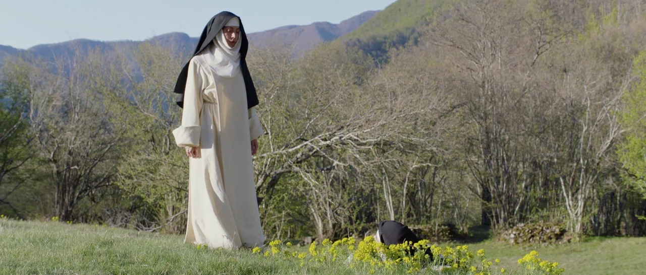 The Little Hours (2017) movie still: fullbody — A green hillside features sparse trees and mountains in the distance under daylight. A nun stands…
