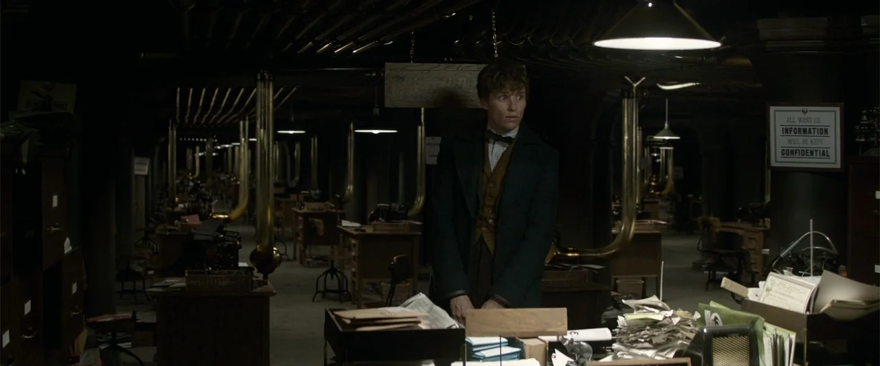Fantastic Beasts and Where to Find Them (2016) movie still: medium — A dimly lit industrial office space features rows of wooden desks and brass…