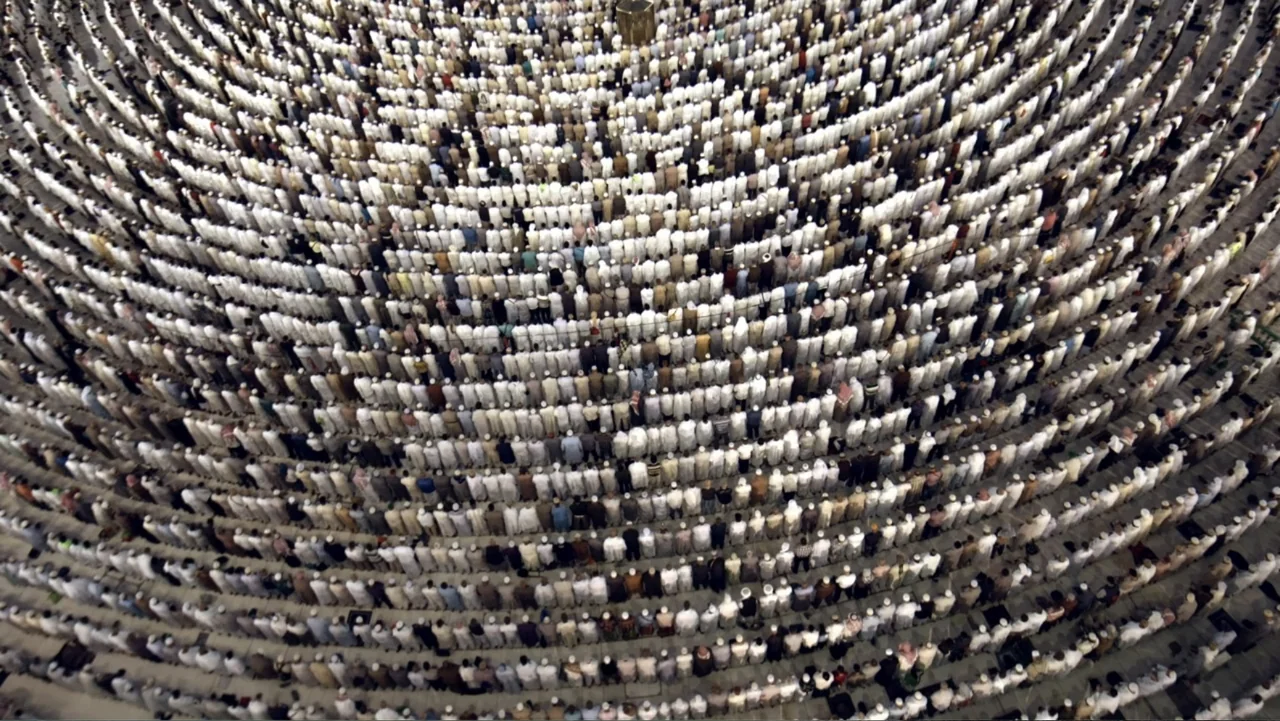 One Day in the Haram (2017) movie still: establishing — A vast stone courtyard filled with a dense crowd of worshippers in white garments. Thousands…