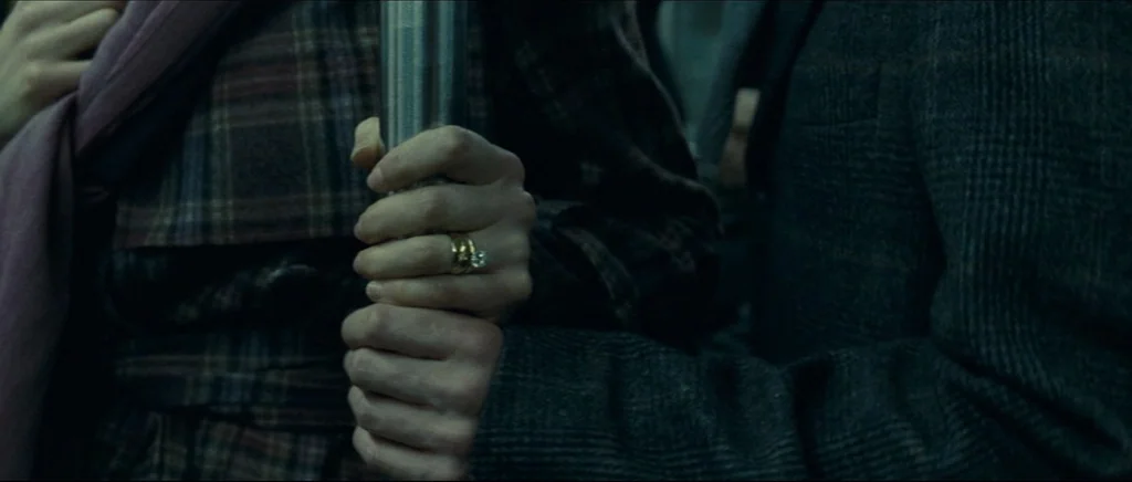 Shame (2011) movie still: closeup — Inside a moving train carriage, hands grip a stainless steel support pole. The hands overlap, displaying a ring…