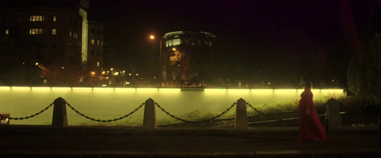 Prevenge (2016) movie still: wide — A woman walks at night along a path backed by a long, illuminated wall. Dark urban structures and trees are…