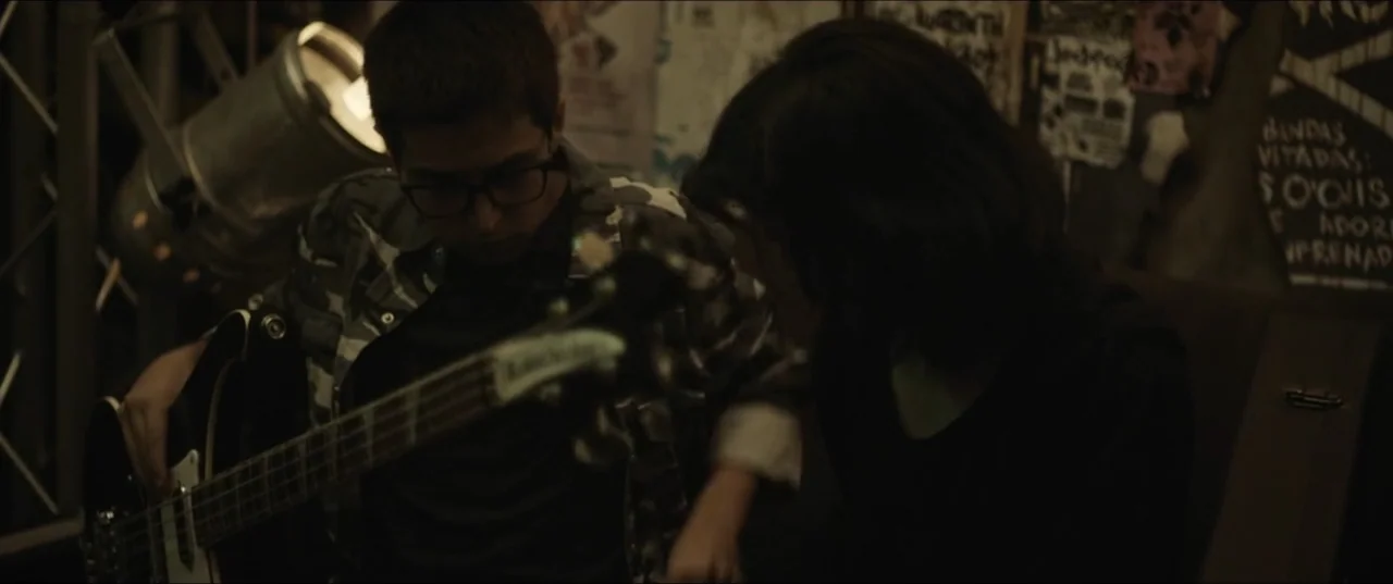 Death to Summer (2021) movie still: medium — A dimly lit room features walls covered in posters and a large lamp fixture. A young man sits holding a…